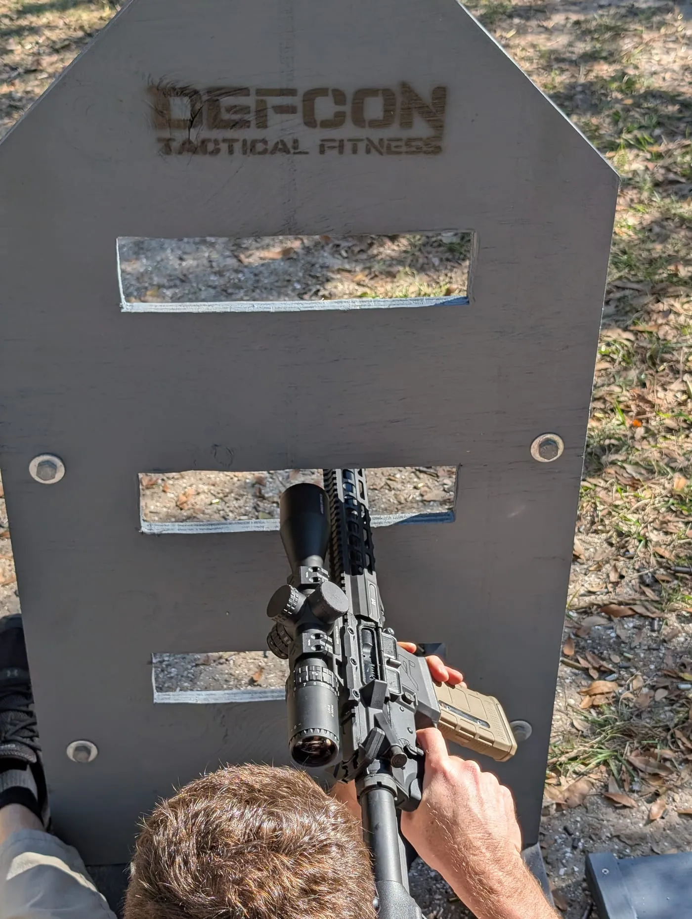 A competitor shooting a rifle through a DEFCON-branded barricade port at a two-gun match